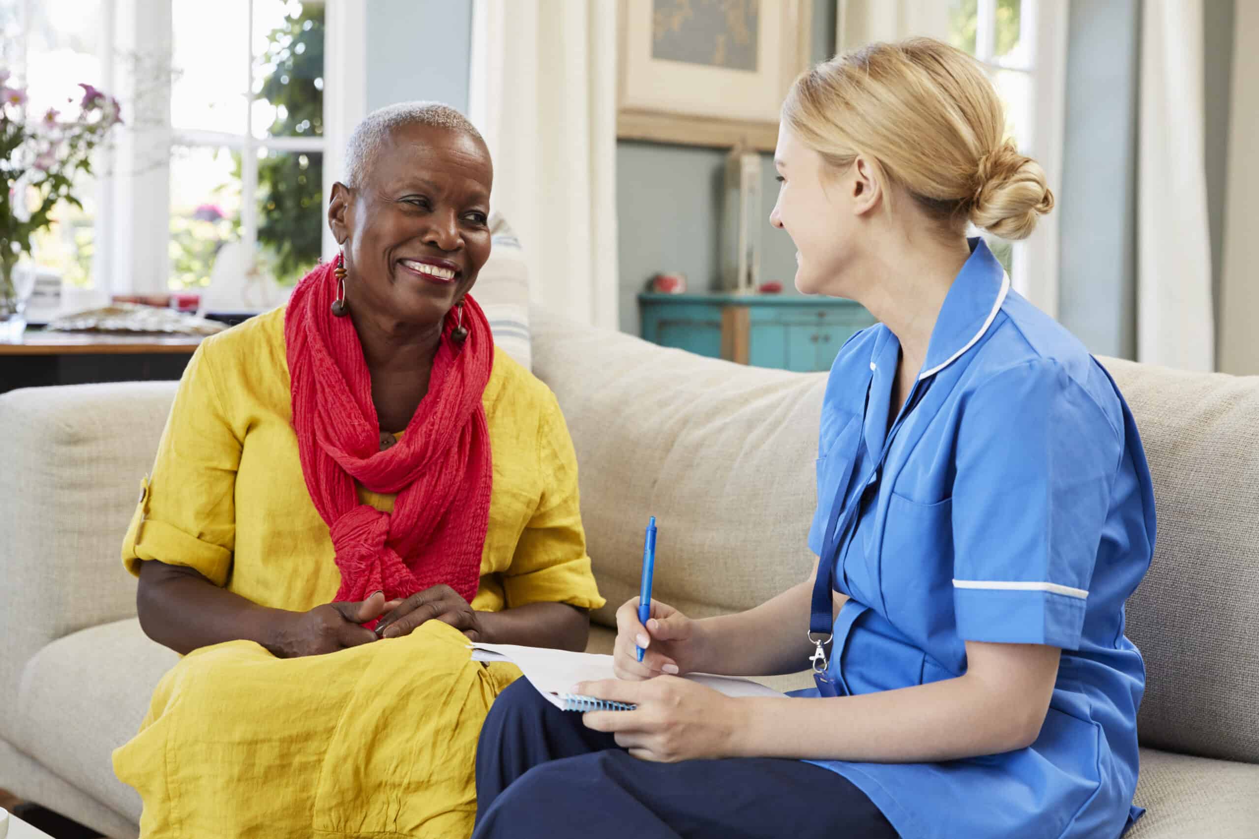An older woman sits on a sofa smiling while talking to a healthcare worker in blue scrubs, who is taking notes during the initial assessment carried out by P4F homecare staff.