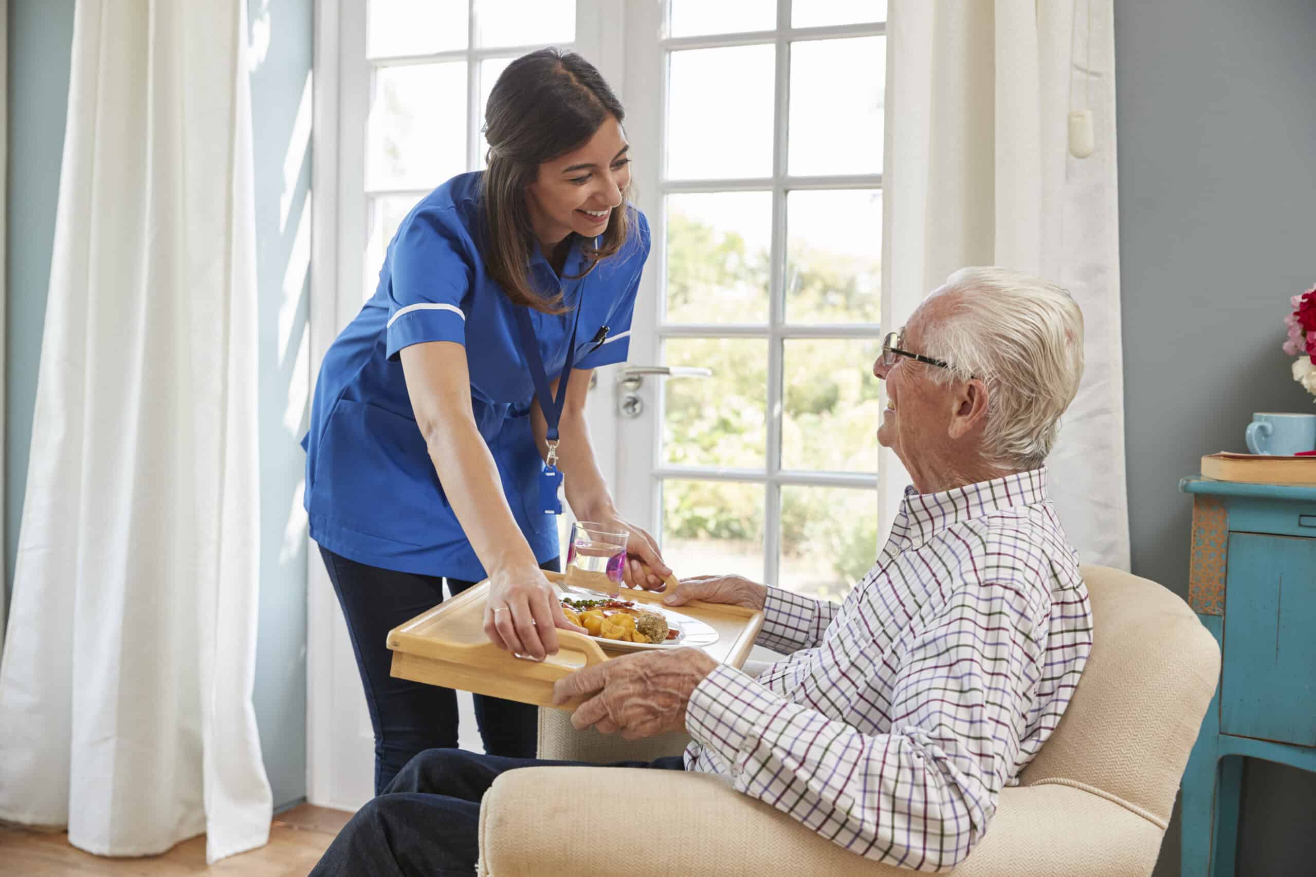 A carer in a blue uniform offers support for daily living activities as she hands a meal tray to an elderly man sitting in a chair by a window in a bright room.