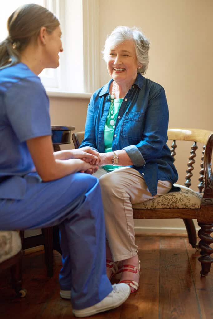 An elderly woman sits and smiles whilst holding hands with a healthcare worker in blue scrubs, enjoying a warm conversation with her P4F Homecare Champion in a well-lit room.